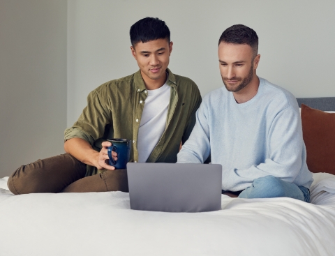 Zwei Männer sitzen entspannt auf einem Bett und schauen auf einen Laptop.